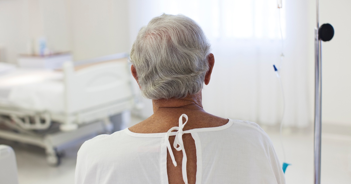 An older patient in a hospital gown sitting on a bed inside a hospital room with medical equipment nearby.