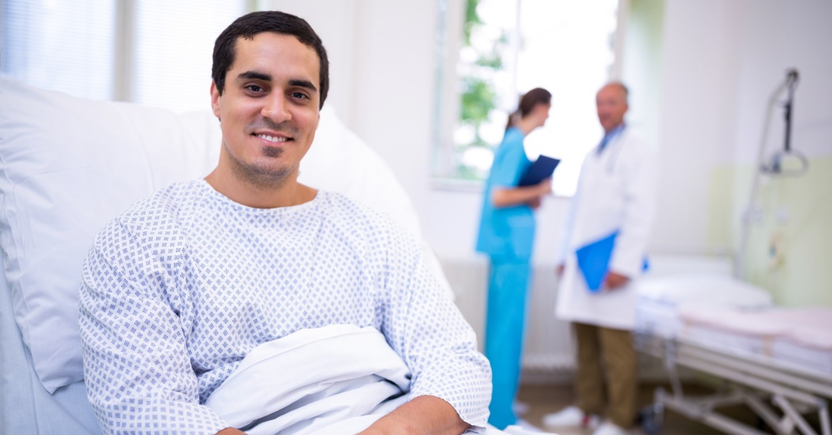 A smiling man in a hospital gown sitting upright in a bed with two medical workers blurred in the background.