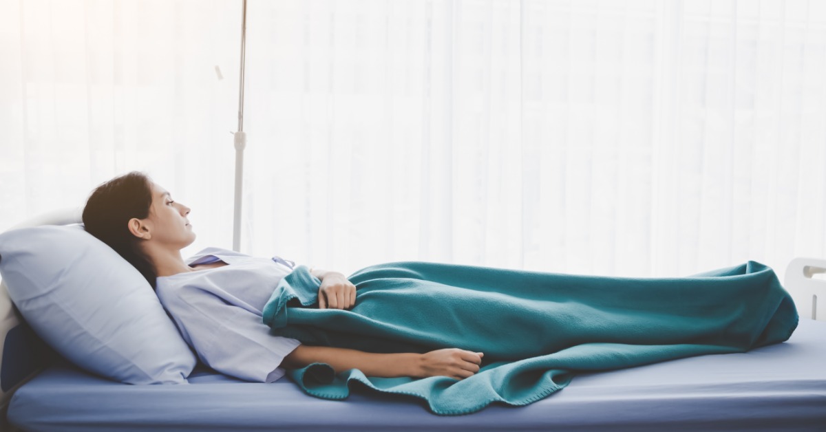 A woman lying in a hospital bed under a teal blanket, with an IV pole beside bright curtains in a white hospital room.