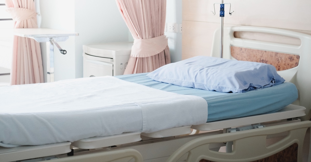 A hospital bed with blue bedding, a white blanket, and a pillow beside pink curtains, an IV pole, and a cabinet.