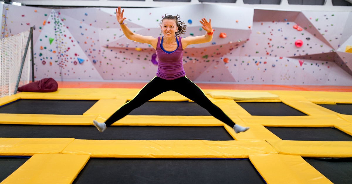 A young person jumping on an indoor trampoline with legs extended in a split while suspended above the surface.