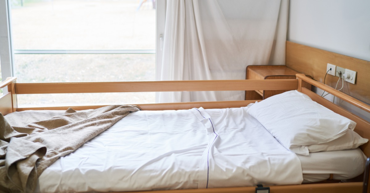 A neatly made empty bed with a folded brown blanket and white pillow near a bright window in a care room.