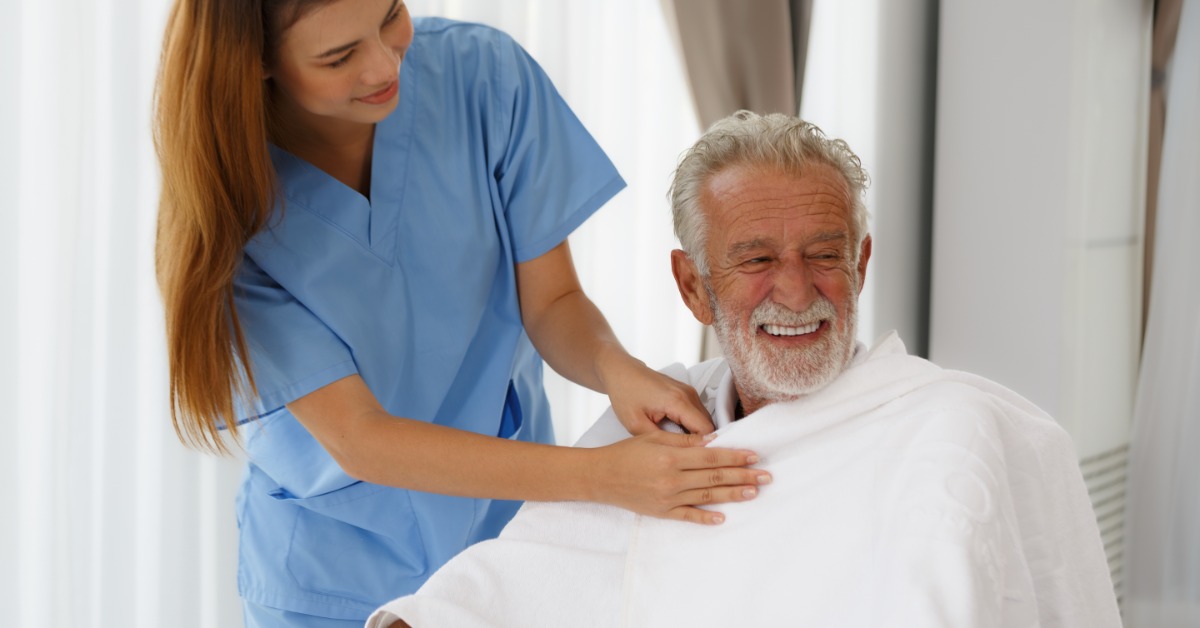 A nurse wearing blue scrubs placing a blanket over a senior man sitting indoors next to a curtained window.