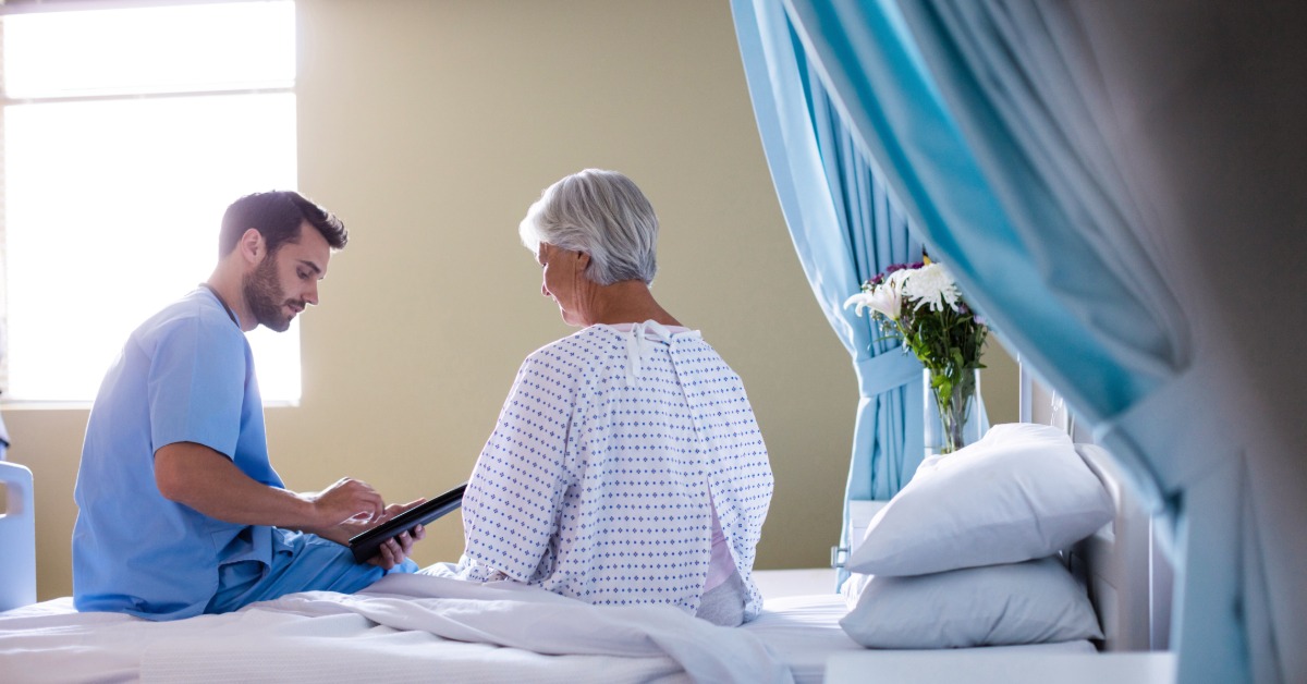 A man in blue scrubs using a tablet beside an older woman sitting on a hospital bed near blue curtains.