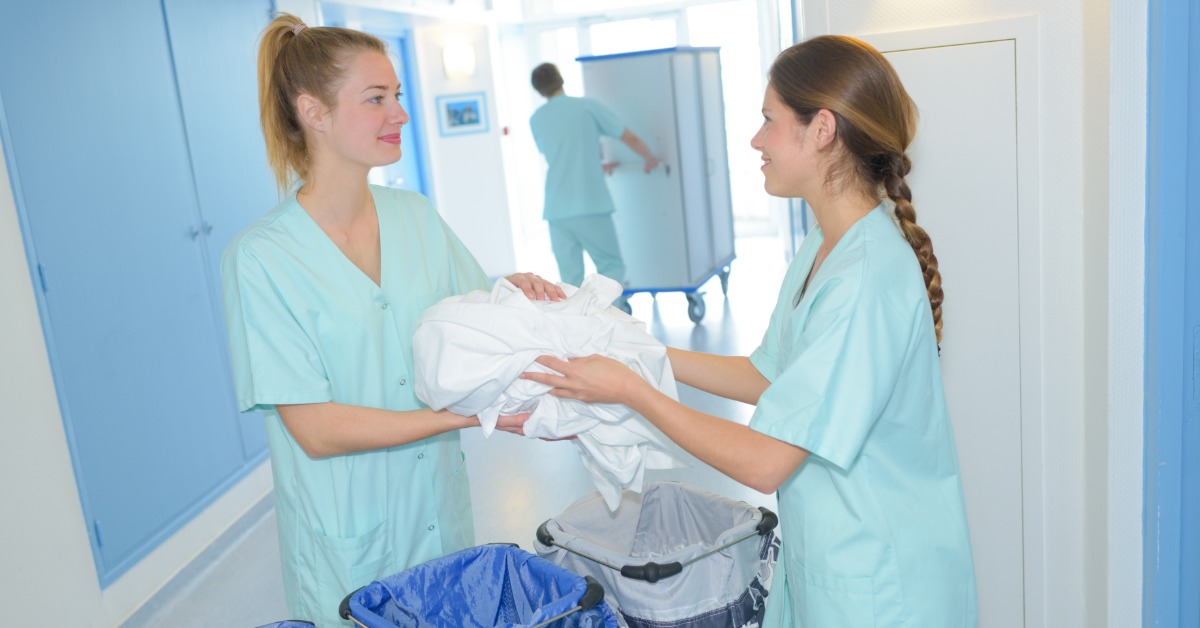 Two women in blue scrubs exchanging white linens beside wheeled laundry carts in a bright hallway with a cart farther back.