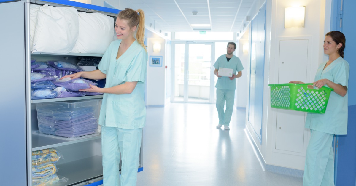 Three people in light blue uniforms organizing wrapped linens and carrying items in a bright hospital hallway.
