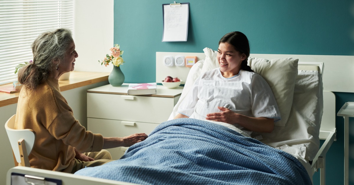 A woman sitting in a hospital bed under a blue blanket, talking with an older woman beside a window.