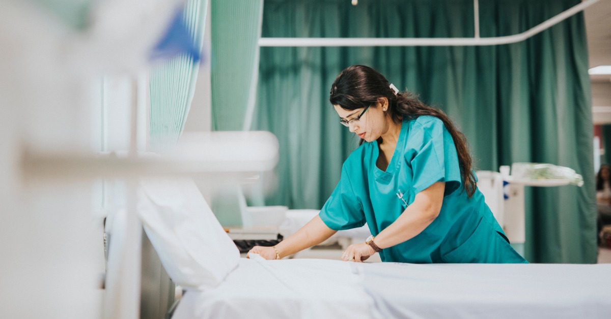 A woman in teal scrubs smoothing a white sheet on a hospital bed beside green privacy curtains inside a hospital room.