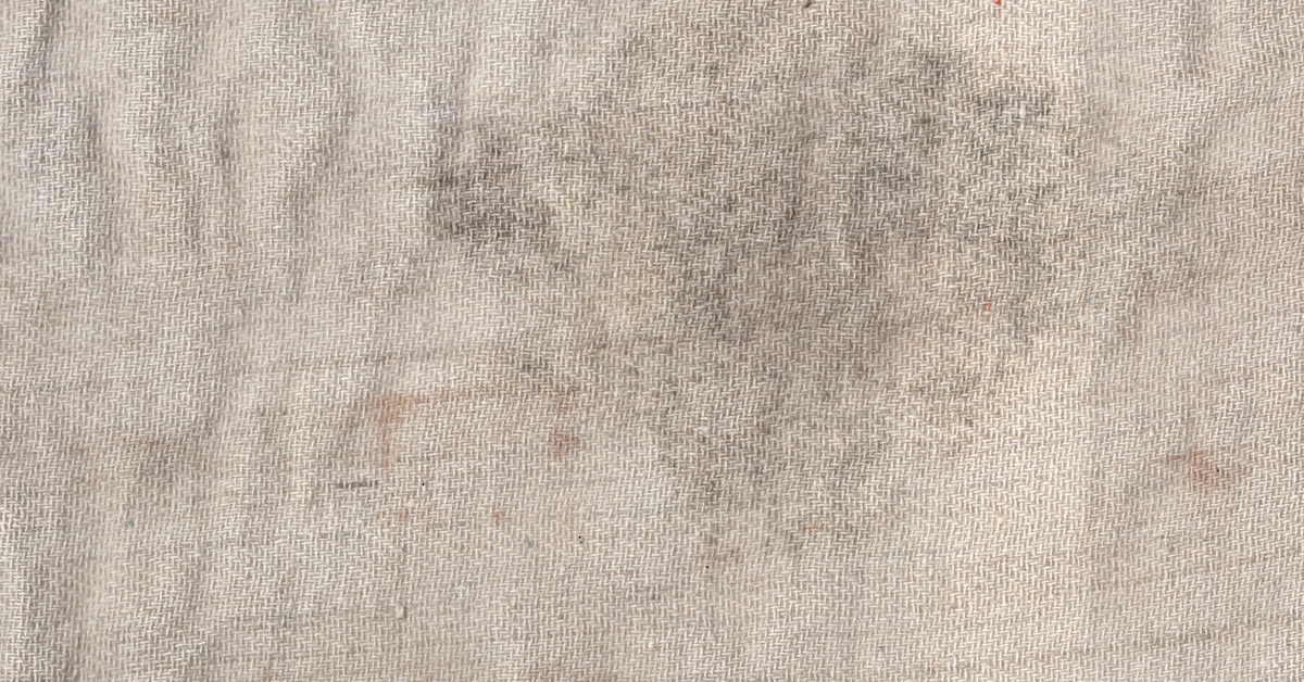 A beige woven fabric surface covered with scattered gray and brown stains, dark smudges, creases, and discoloration.