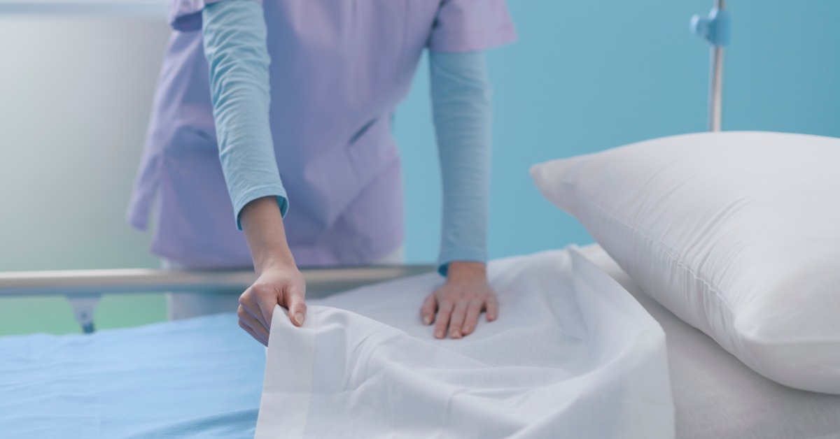 A nurse smoothing a clean, white bed sheet over a hospital bed while standing beside it in a medical room.