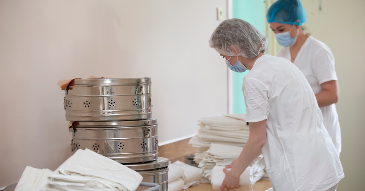Two women wearing scrubs, masks, and hair nets folding white sheets on a wooden table in a hospital laundry room.