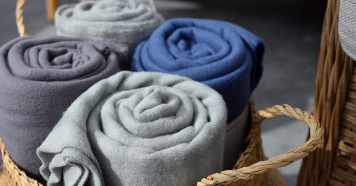 A wicker basket holding rolled fleece blankets in different colors, next to a wicker chair with an off-white cushion.