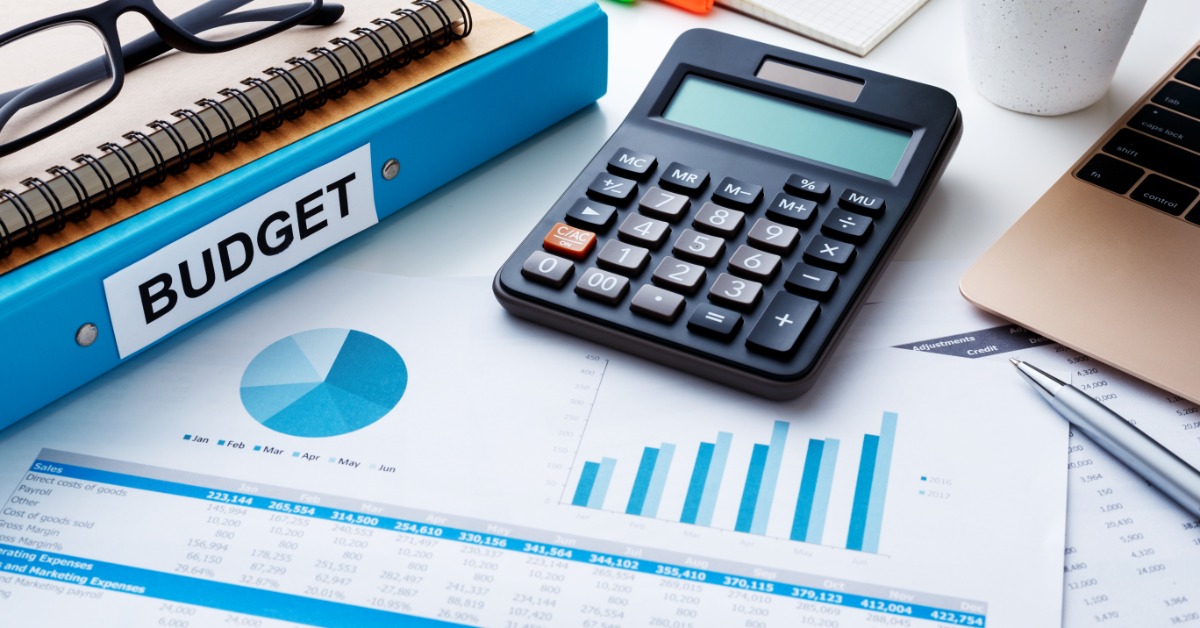 A financial chart report with printed graphs, a calculator, a pen, and a laptop arranged on a work desk.
