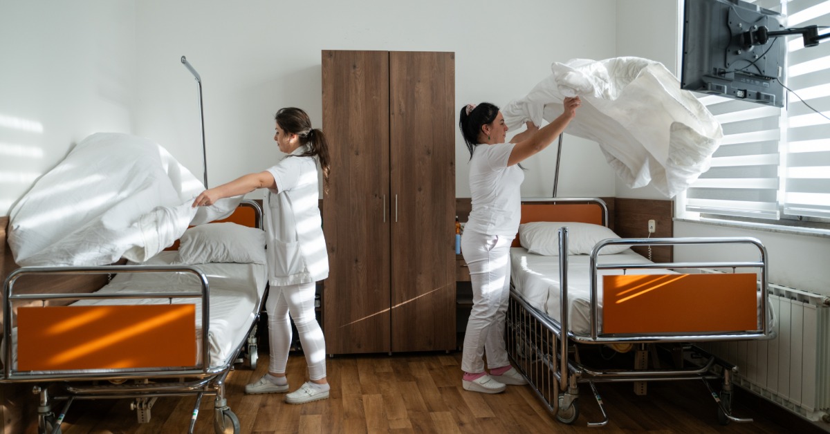 Two women wearing white uniforms changing linens on adjacent beds in a clean room with bright windows.