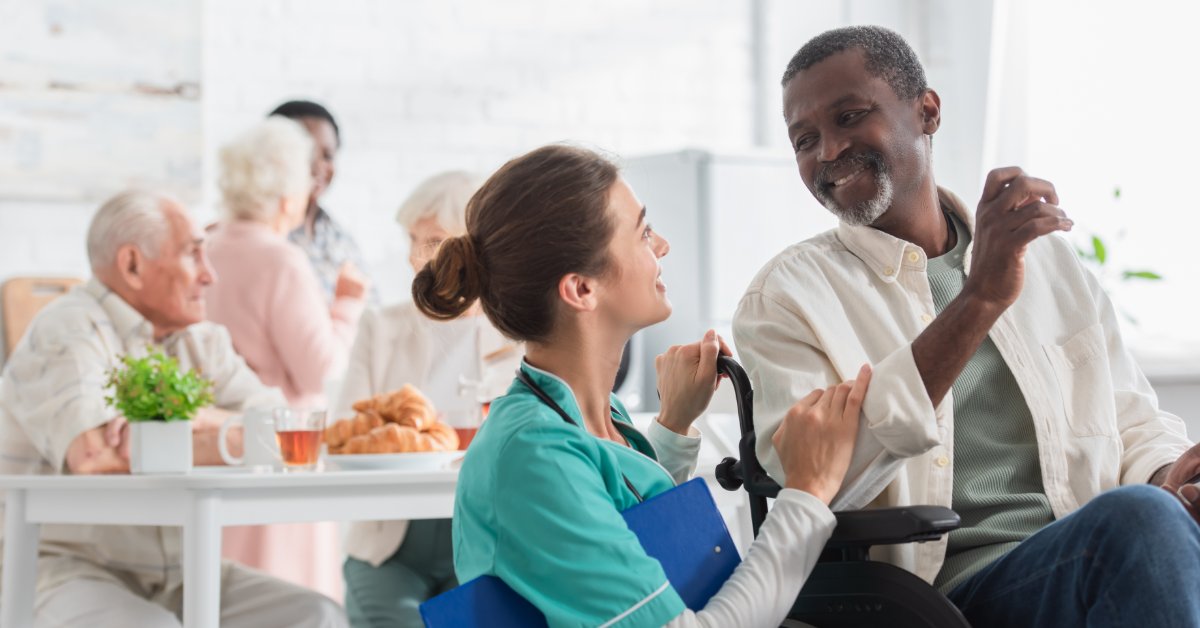 A woman in teal scrubs holding a clipboard while talking with a smiling man seated in a wheelchair near a table.