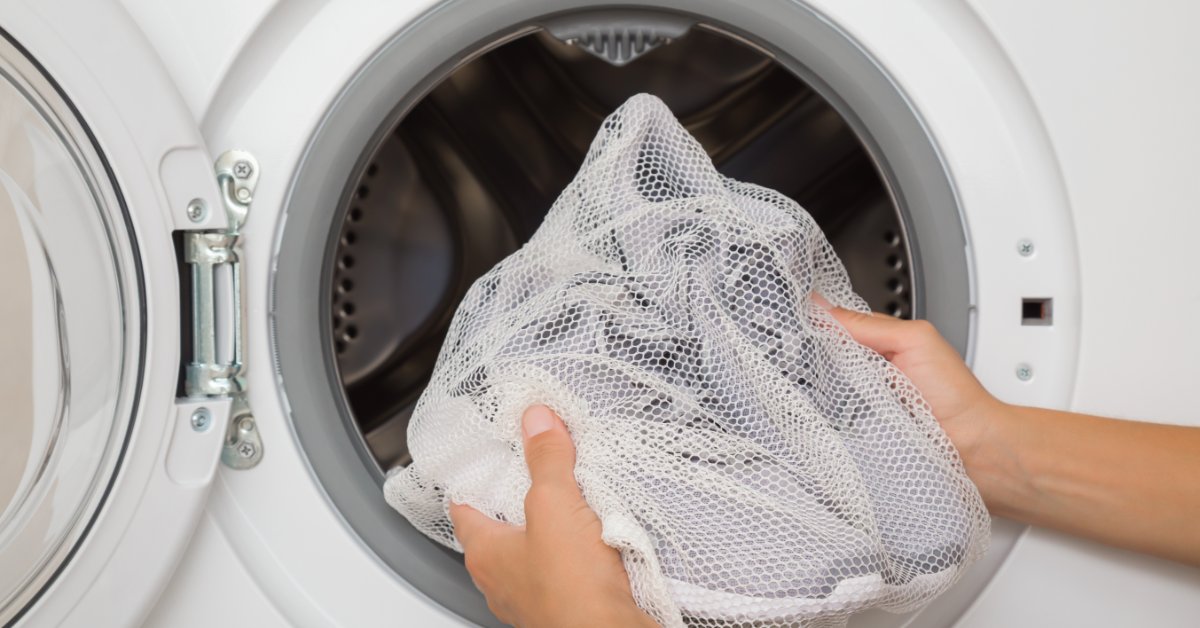 Hands holding a white mesh bag of athletic shoes in front of an open white washing machine door and round metal drum.