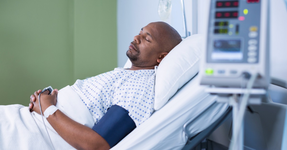 A man resting in a hospital bed with a blood pressure cuff, a handheld device, and blurred medical equipment.