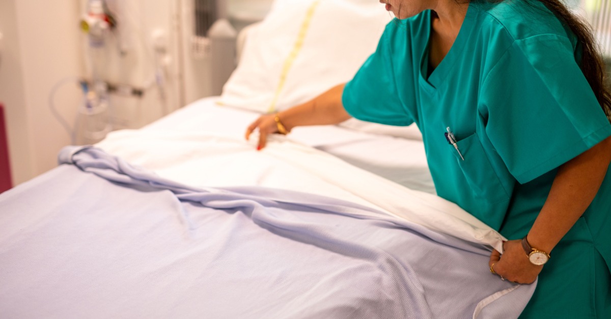 A person in teal scrubs adjusting white and blue bedding on a hospital bed inside a bright patient room.