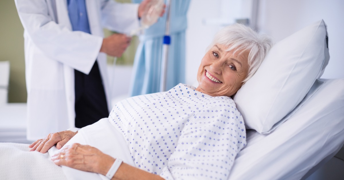 An older woman smiling while lying in a raised bed as a person in a white coat adjusts an IV line nearby.