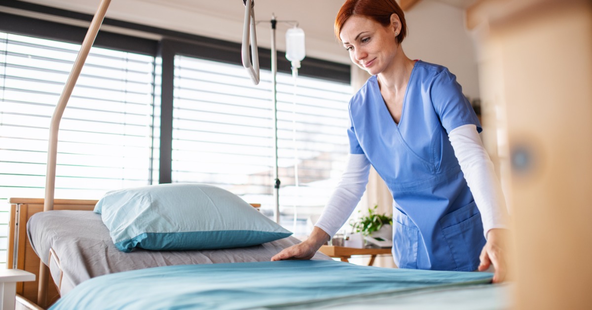 A woman in blue scrubs smoothing a turquoise blanket on an empty bed near windows and medical equipment.