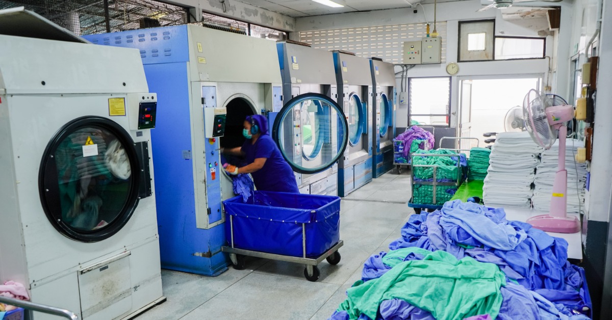 A person wearing a mask loading blue fabric into industrial laundry machines near piles of colored cloths.